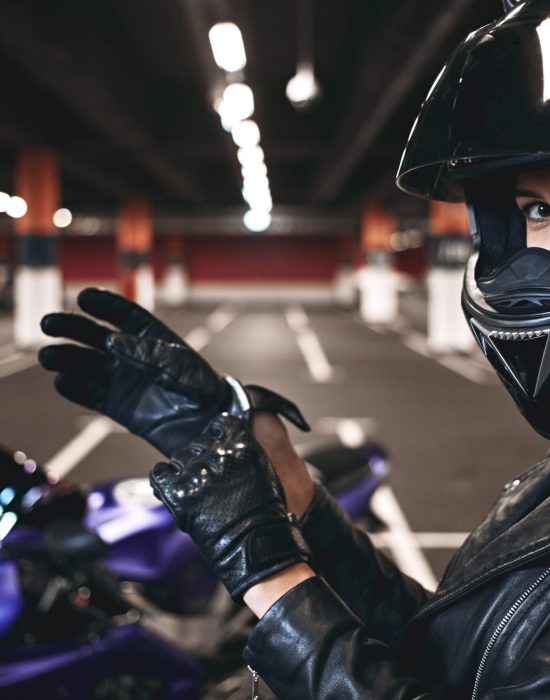 People, urban lifestyle, extreme sports and adrenaline conept. Sideways portrait of palyful styligh young Caucasian motorcycle rider in fashionable black leather jacket and helmet, adjusting gloves